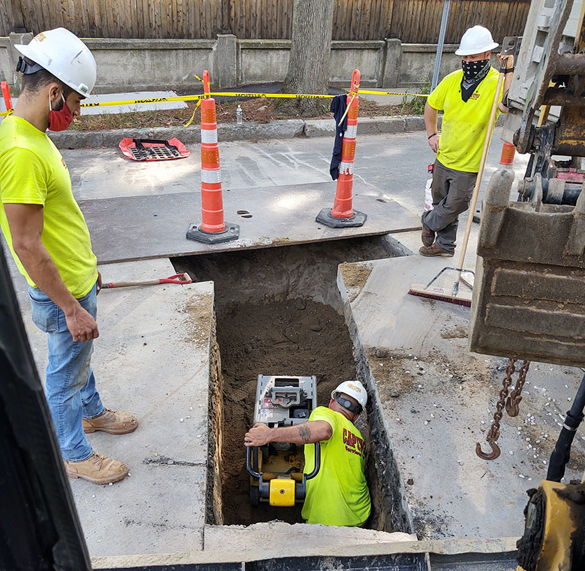 2 underground utilities workers standing above and looking down at a thrid contractor in utilities hole.